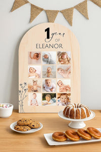 First birthday sign with 12-month baby photo collage leaning against wall on dessert table with bundt cake, cookies, and doughnuts