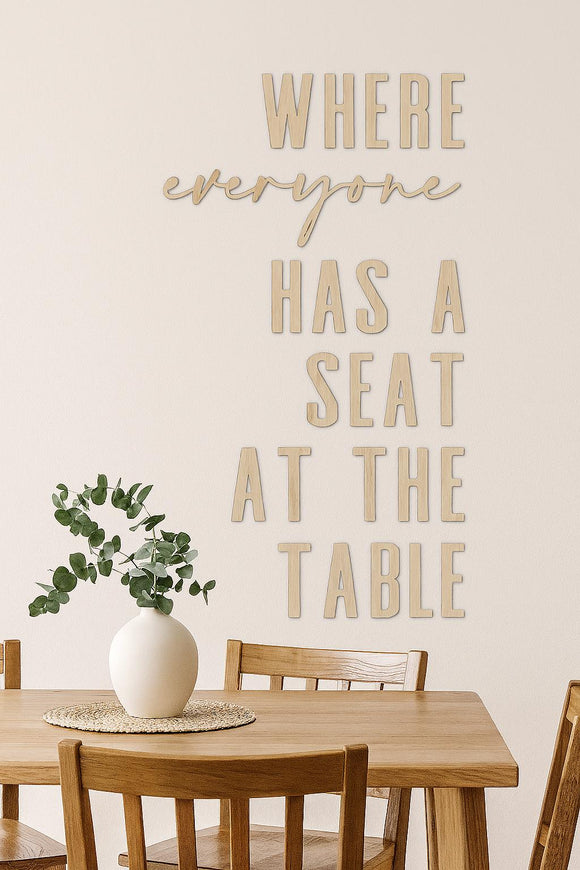 Laser cut wood wall sign reading "Where everyone has a seat at the table" displayed on a neutral dining room wall with wooden table and chairs beneath.